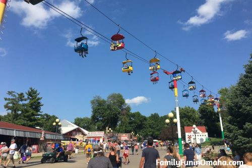 A group of people walking on the grounds of the Iowa State Fair with sky ride chairs overhead.