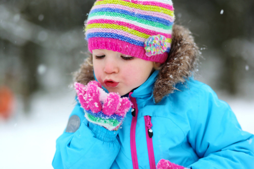 A little girl wearing a hat and mittens blowing snow off her hands